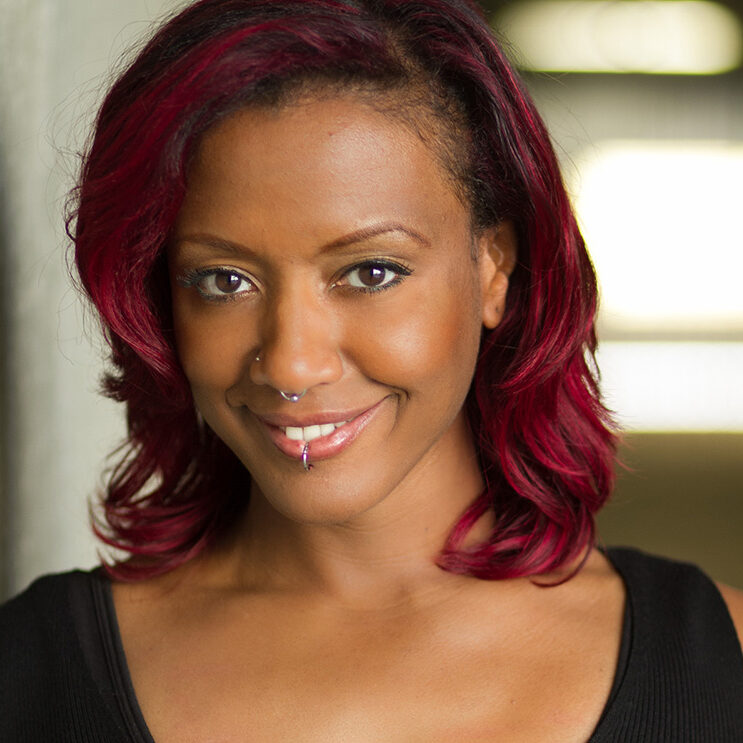 A person with shoulder-length red hair and a facial piercing smiles at the camera, their black top subtly contrasting the softly blurred background. The warmth in their expression mirrors the spirit of community support at the Los Angeles Regional Food Bank.
