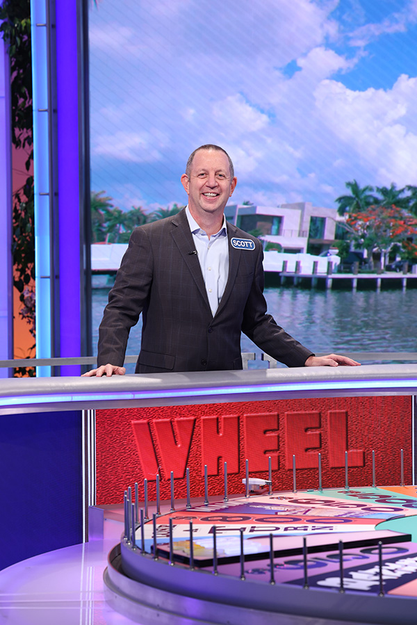A contestant stands smiling behind a game show podium labeled "WHEEL," proudly wearing a name tag supporting the Los Angeles Regional Food Bank. The background features a waterfront scene with modern buildings and palm trees, complementing his sharp suit.
