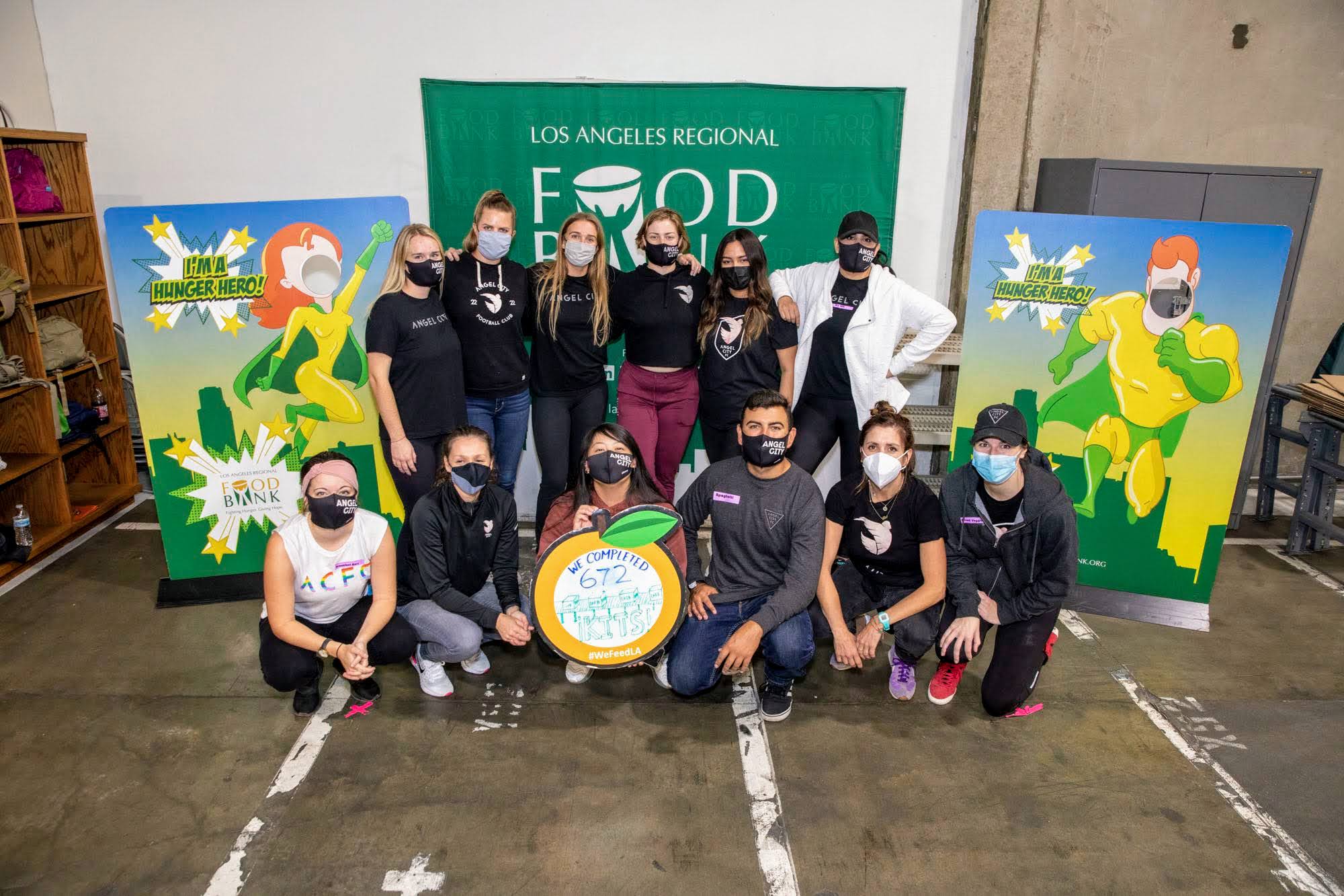 At the Los Angeles Regional Food Bank, a group of masked individuals poses indoors holding a sign with "672," surrounded by vibrant superhero-themed posters, capturing the warehouse setting's dynamic spirit.