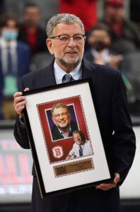 A man in glasses and a suit proudly holds a framed picture and plaque from the Los Angeles Regional Food Bank. The frame showcases two photos of him and an engraved plate with text. Behind him, a blurred indoor setting reveals bustling people.