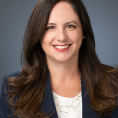 A woman with long brown hair and a bright smile is wearing a dark blazer over a white top, volunteering passionately at the Los Angeles Regional Food Bank. The background is a plain gray color.