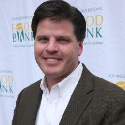 A person in a white shirt and dark jacket smiles at the camera, standing in front of a backdrop with "Los Angeles Regional Food Bank" written on it.