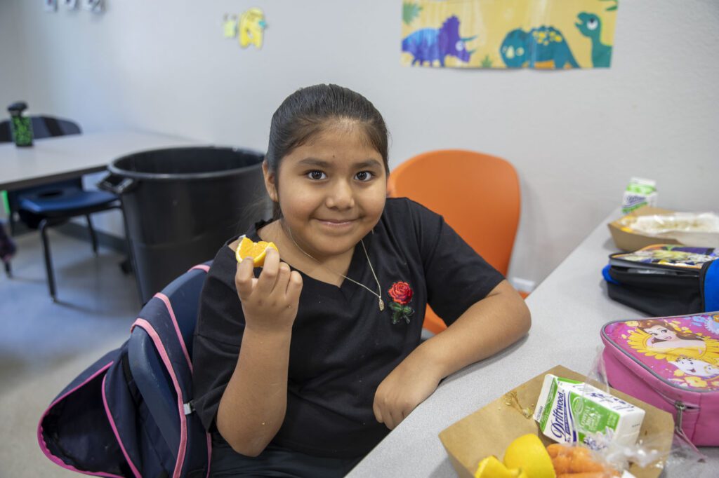 A young girl is holding fruit as part of her lunch during a free summer lunch distribution at Koreatown Youth and Community Center in the summer of 2023