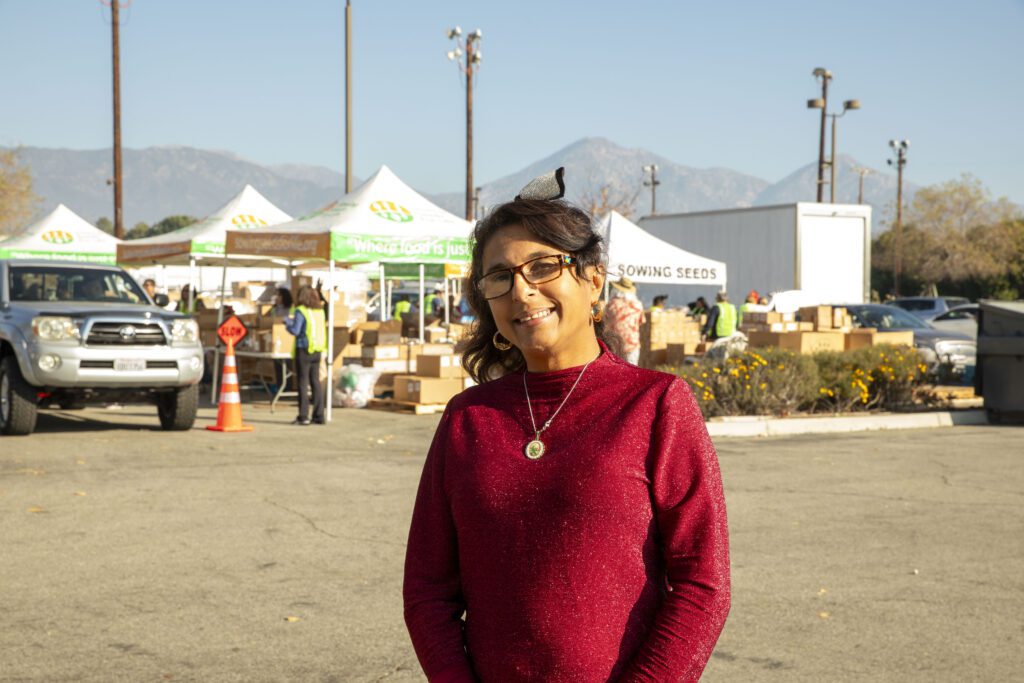 A person wearing glasses and a red sweater smiles at the camera. In the background, volunteers from the Los Angeles Regional Food Bank are near tents and boxes, possibly distributing goods. The setting is outdoors with mountains in the distance and a parked car nearby.