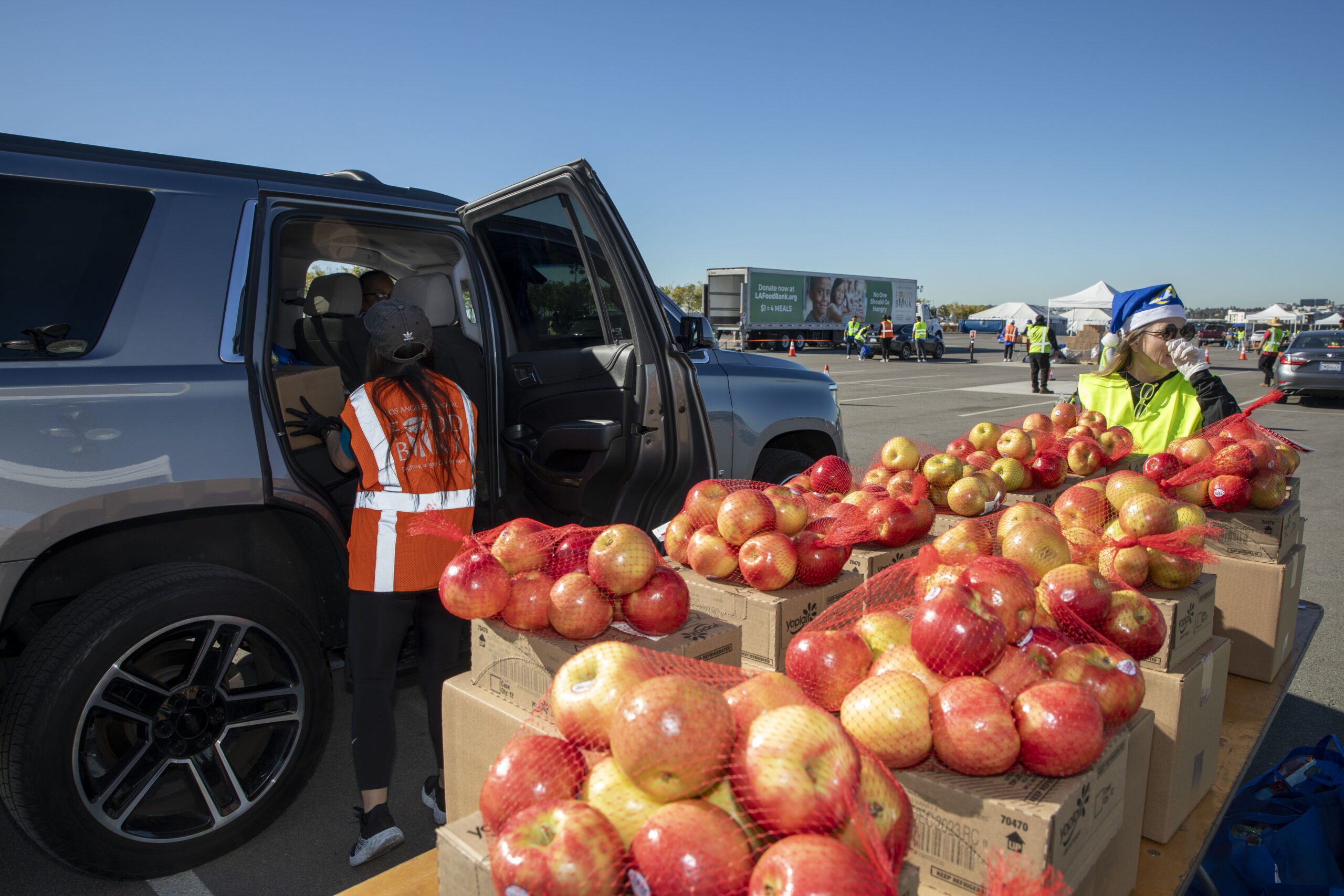 231205-Rams-SoFi-Distribution-9583 Volunteers from the Los Angeles Regional Food Bank distribute bags of apples to people in cars at a bustling food donation event. Tables are lined with boxes of fresh apples against the backdrop of a clear, blue sky.