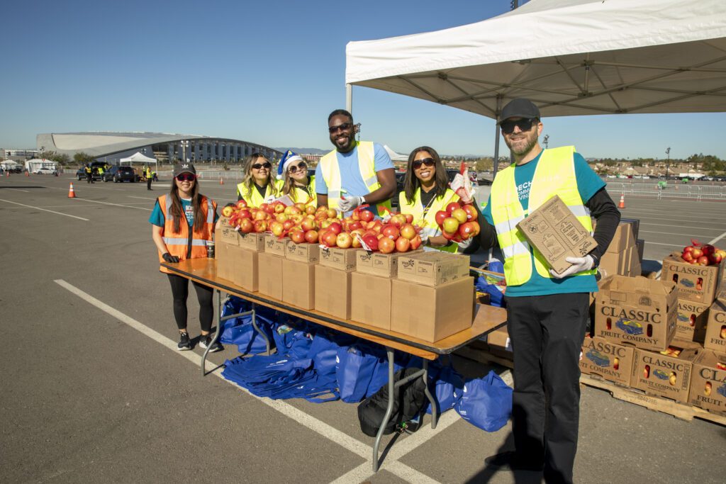 A group of people in safety vests gather around a table with boxes of apples at an outdoor Los Angeles Regional Food Bank event. They are smiling under a white canopy, with the open space and a large building visible in the distance.