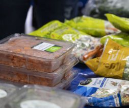 A table with various packaged foods from the Los Angeles Regional Food Bank, including pre-cut vegetables and salad mixes. Some packages are stacked and labeled, displayed against a blue cloth backdrop.