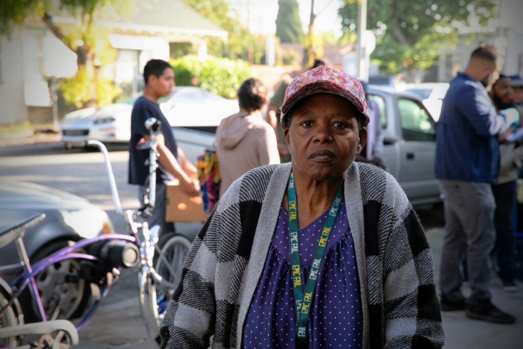 An older woman in a floral cap and striped sweater stands on a sidewalk with her Los Angeles Regional Food Bank lanyard around her neck. In the background, people mingle near a street bustling with cars and bicycles.