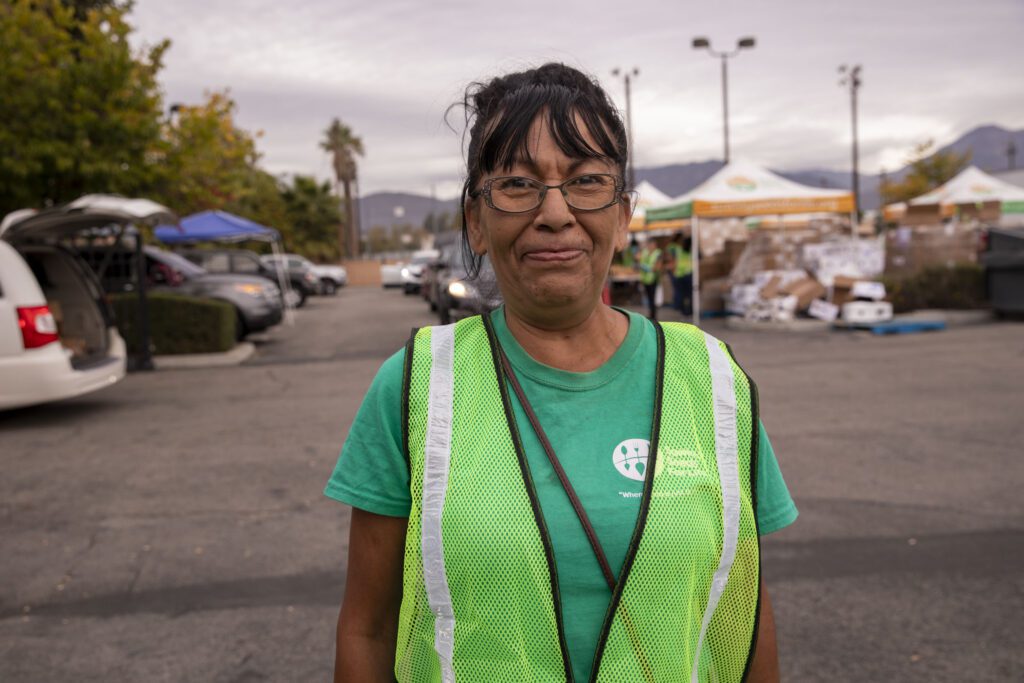 A woman wearing glasses, a green safety vest, and a green shirt stands in a parking lot at the Los Angeles Regional Food Bank. Tents and cars are visible in the background, framed by trees and mountains under a cloudy sky.