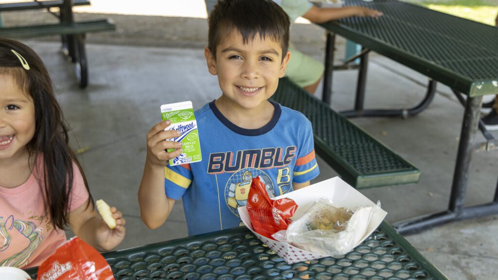 A young boy, sitting at a picnic table, smiles while holding a carton of milk from the Los Angeles Regional Food Bank. He is wearing a blue t-shirt with colorful graphics. Next to him, another child happily enjoys food, including wrapped sandwiches and a cookie.