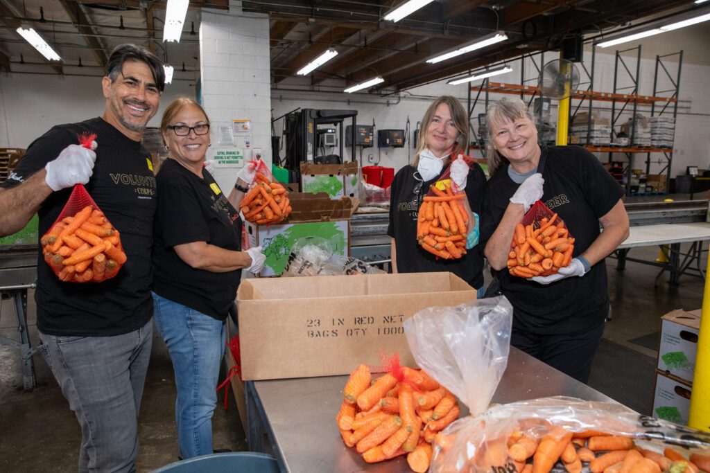 Four volunteers happily hold bags of carrots in a bustling Los Angeles Regional Food Bank warehouse, surrounded by boxes and a large bag of carrots on a table. Wearing gloves and black 