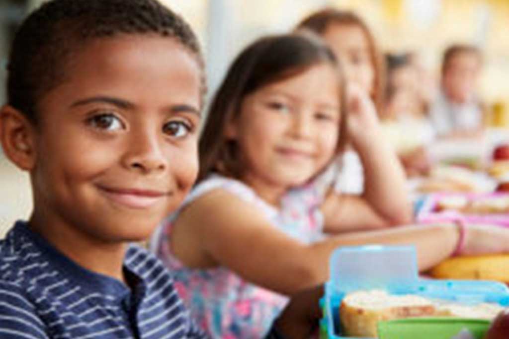 LAFB-Ways-to-give-Monthly-Giving Two children sit at a table, grinning with lunch boxes from the Los Angeles Regional Food Bank before them. Other kids are blurred in the background, but the spotlight is on the boy with curly hair and his cheerful companion beside him.