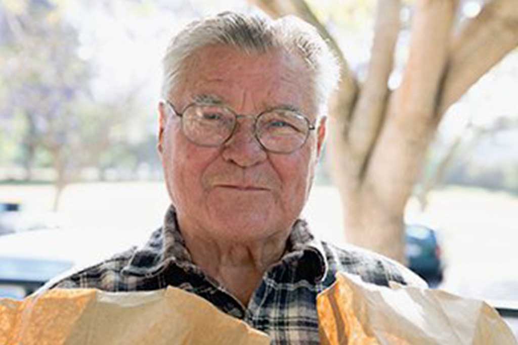 LAFB-Ways-to-give-planned-giving An elderly man with glasses and a plaid shirt stands outdoors clutching a brown paper bag, which he recently received from the Los Angeles Regional Food Bank. The background showcases blurred trees under bright sunlight, highlighting the warmth of community support.