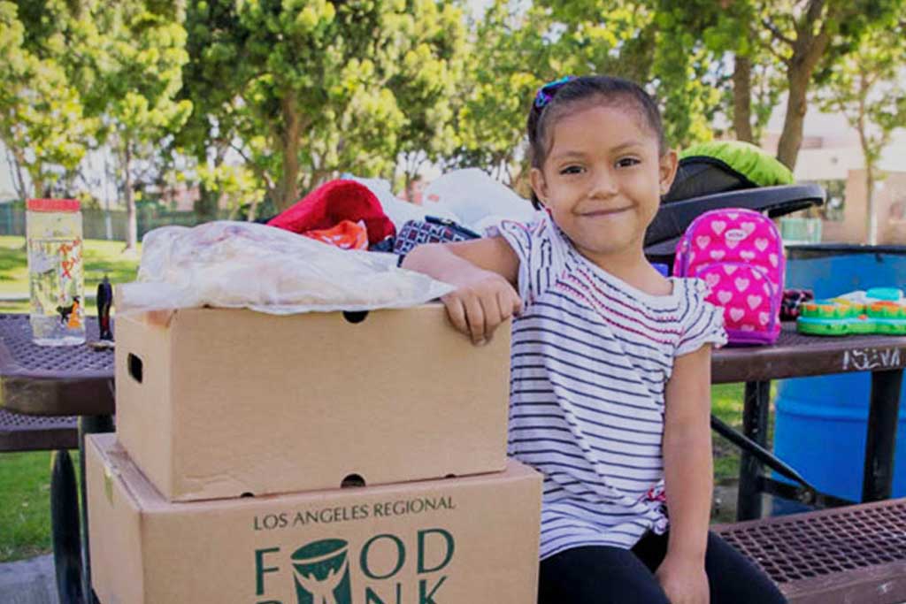 LAFB-Ways-to-give-Cryptocurrency A young girl smiles while leaning on a stack of food boxes from the Los Angeles Regional Food Bank at a park. She is wearing a striped shirt with a pink hair bow, and there is a colorful backpack and trees in the background.