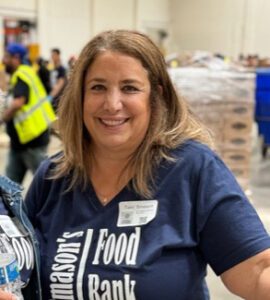 A woman with long hair wearing a blue shirt smiles in a bustling warehouse environment at the Los Angeles Regional Food Bank. People in safety vests and boxes of supplies fill the background, creating an atmosphere that suggests a vibrant volunteer or community event.