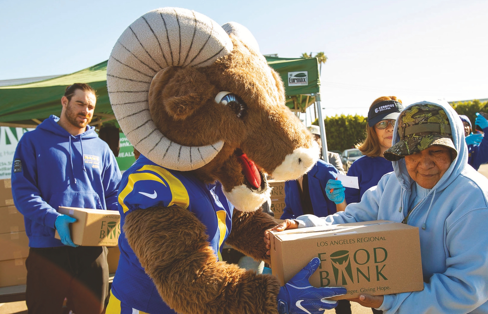 In front of a green tent, a person in a Ram mascot costume hands a Los Angeles Regional Food Bank box to an elderly person wearing a blue hoodie and camouflage cap. Other volunteers in blue shirts assist enthusiastically in the background.
