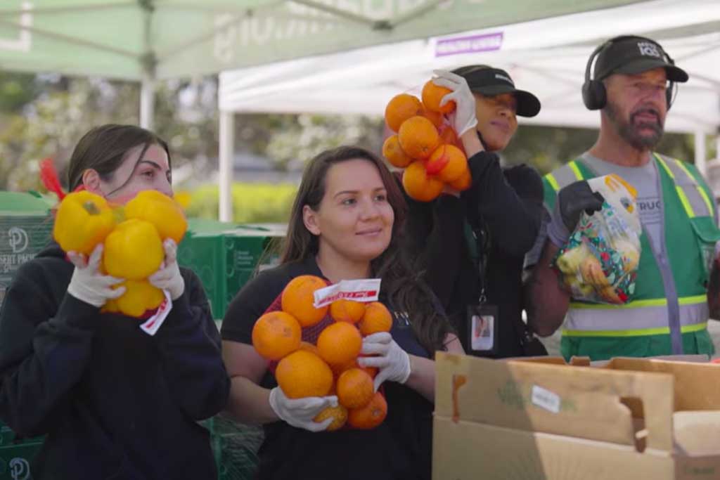 LAFB-Ways-to-give-Roth-IRA Four people under a tent hold fresh produce: two with oranges, one with bell peppers, and another with a bag of mixed fruits and vegetables. They stand behind a table with boxes, wearing gloves and casual clothes, supporting the Los Angeles Regional Food Bank initiative.