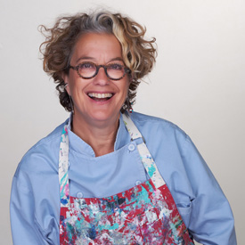 Susan-Feniger-27-275 A person with curly hair and glasses is smiling, wearing a light blue shirt and a colorful, paint-splattered apron against a plain background, as they volunteer their art skills for the Los Angeles Regional Food Bank.