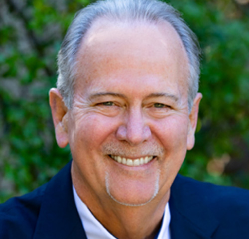 A smiling man with short gray hair and a goatee, donned in a dark suit jacket, stands outdoors against a blurred green backdrop. His commitment to the Los Angeles Regional Food Bank shines through his warm demeanor.