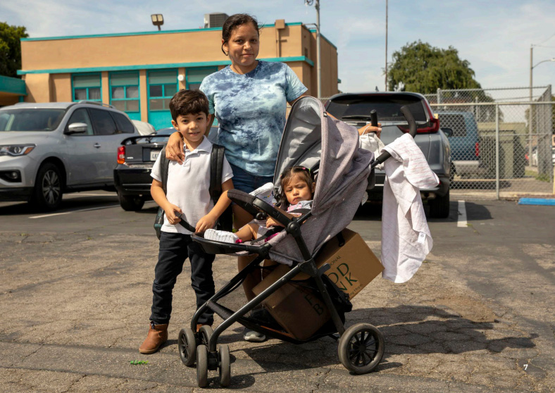 A woman stands beside a young boy and a toddler in a stroller outside the Los Angeles Regional Food Bank. They are in a parking lot with cars and a building behind them. The toddler holds a box in the stroller's basket, while various items hang from the handle.