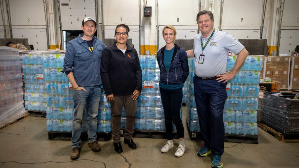 Four people stand in a warehouse setting in front of stacks of bottled water. Three are casually dressed; the fourth is in a polo shirt and dark pants. They are smiling and appear to be part of a community or volunteer effort.