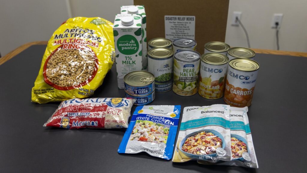 A collection of food items includes a bag of cereal, cartons of milk, canned vegetables, canned tuna, instant noodles, microwaveable rice, and a box labeled 