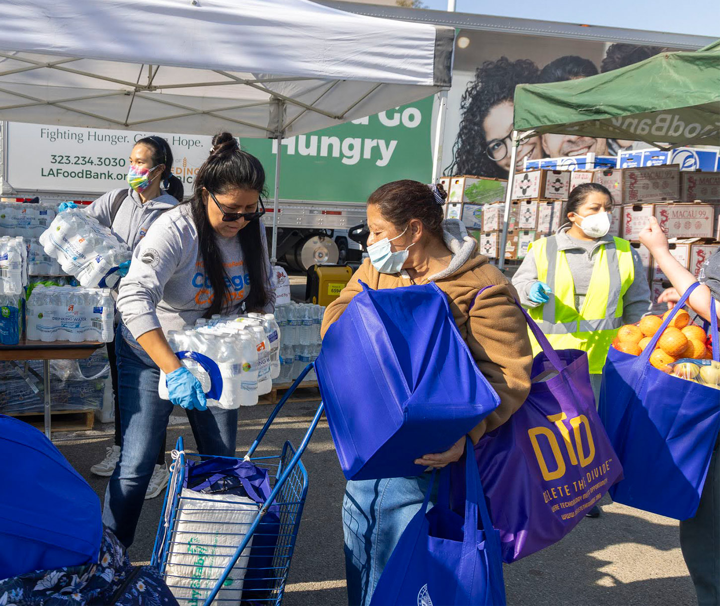 fire-disaster-relief-los-angeles-regional-food-bank Volunteers distribute food and water at a community event aimed at earthquake relief. A woman places bottled water in a cart, while another in a brown jacket holds a blue bag. People wear masks, and a truck with "Fighting Hunger" is visible. Bagged apples are nearby.