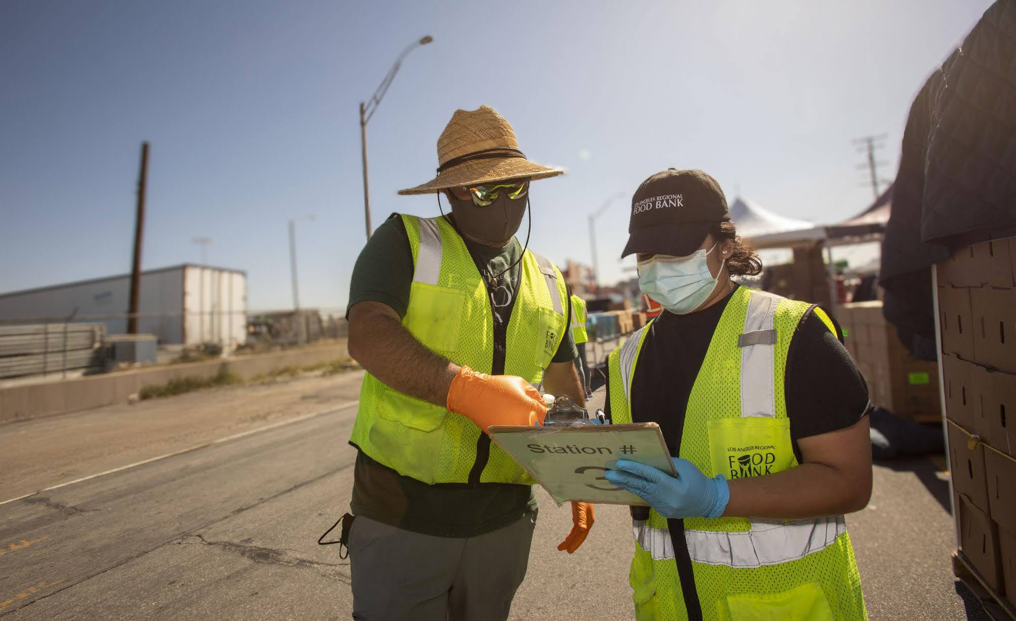210327-Donated-Masks-9040 (1) Two people in safety vests and masks stand outdoors amid earthquake relief efforts. One, in a wide-brimmed hat, gestures at a clipboard held by the other, who sports a baseball cap. They stand next to pallets at what appears to be a bustling distribution site.