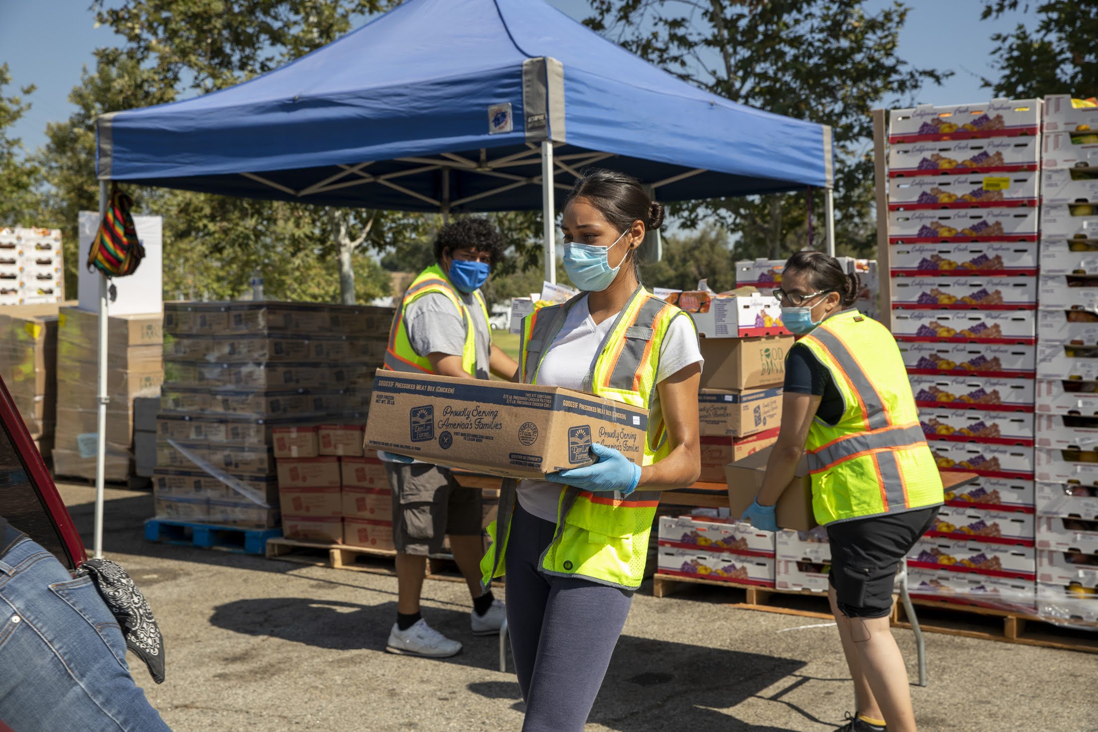 200722-Roger-Mission-Hills-5018 Volunteers wearing masks and safety vests distribute boxes of food at an outdoor event to support earthquake victims. A woman in the foreground carries a box, while others work nearby. Stacks of food boxes and a blue canopy are visible in the background.