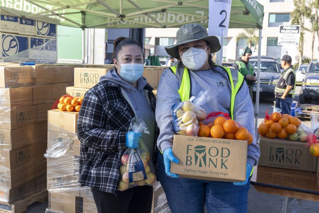 Two people wearing masks stand outdoors at a food bank under a green tent. They hold bags and a box filled with fresh produce, including oranges and apples. Stacks of boxes and a 