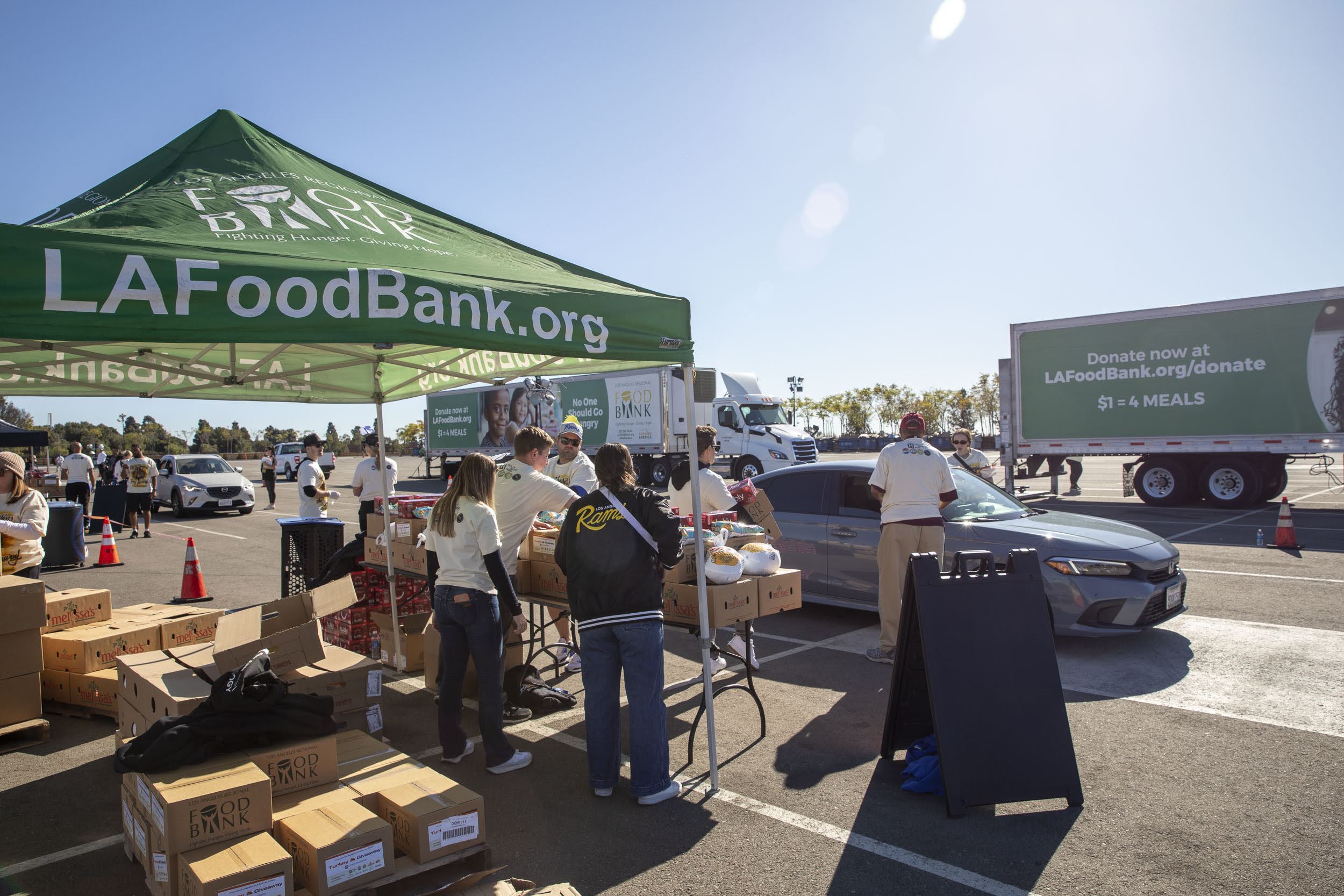 Volunteers distribute food at an outdoor drive-thru event, showcasing their dedication in an inspiring Impact Report. A green tent with "LAFoodBank.org" stands beside a large truck as people load boxes into cars under a clear blue sky.