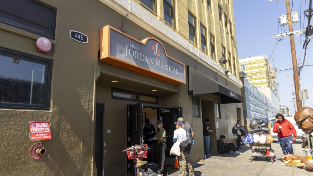 People gather outside the entrance of Fred Jordan Missions on a bustling city street, where hope intertwines with need. A few individuals are seated on the sidewalk beside a building that echoes stories of food pantries and community support. Nearby, someone pushes a cart filled with belongings.