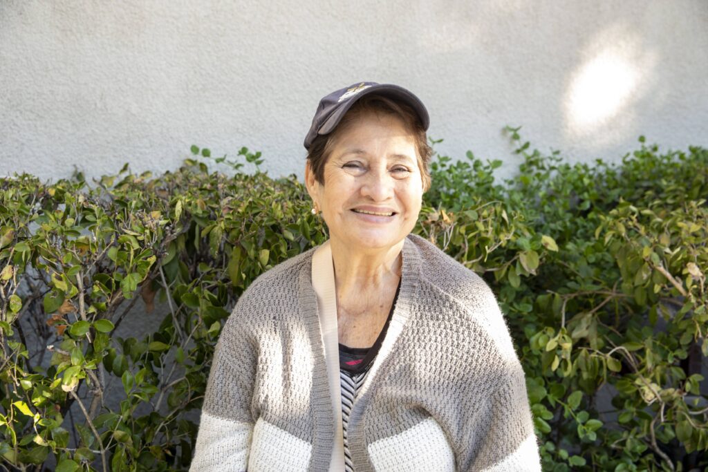 Elderly woman smiling warmly, wearing a knitted sweater and cap, sitting in front of a leafy hedge with a neutral wall in the background, sharing stories of her beloved community food bank.