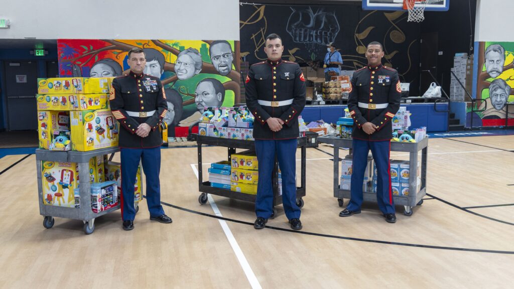 Three individuals in military uniforms stand beside carts filled with boxed toys in a gym, reminiscent of a bustling food bank. Behind them, colorful murals of diverse faces are painted on the walls, adding vibrancy to the scene.