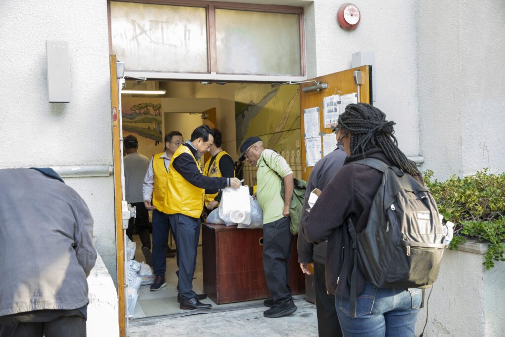 At the community center entrance, a food bank volunteer in a yellow vest hands a bag to an older man, while others wait patiently in line. The lively scene unfolds outdoors, against the backdrop of a colorful mural inside the building.