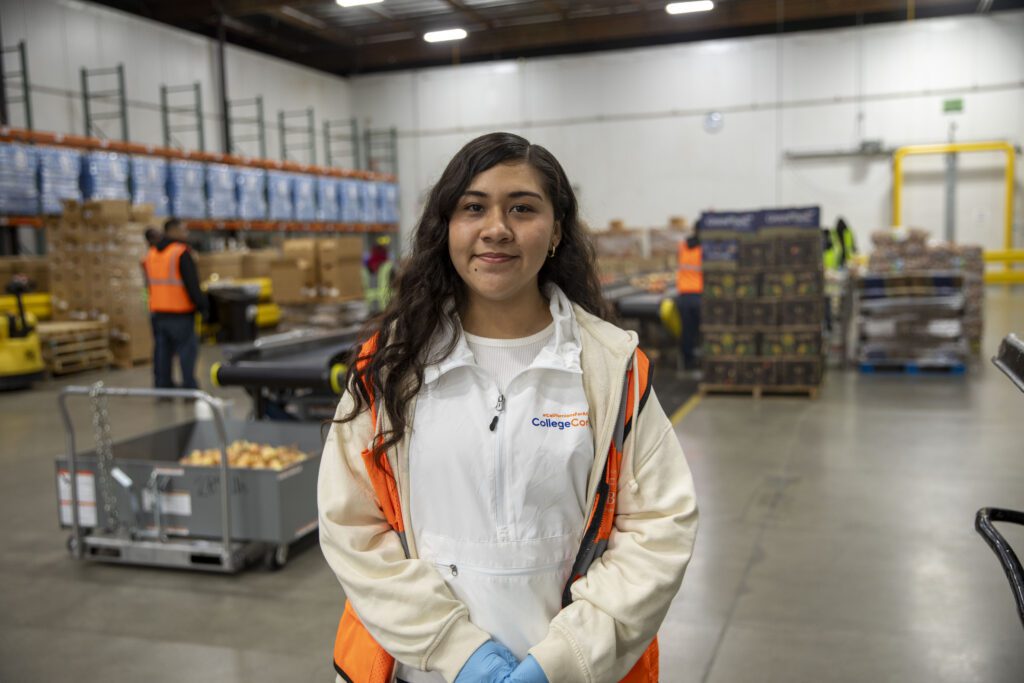 A young woman wearing a white hoodie, orange safety vest, and gloves stands smiling in a warehouse where shelves hold boxes of food from the bar, with other workers busy in the background.