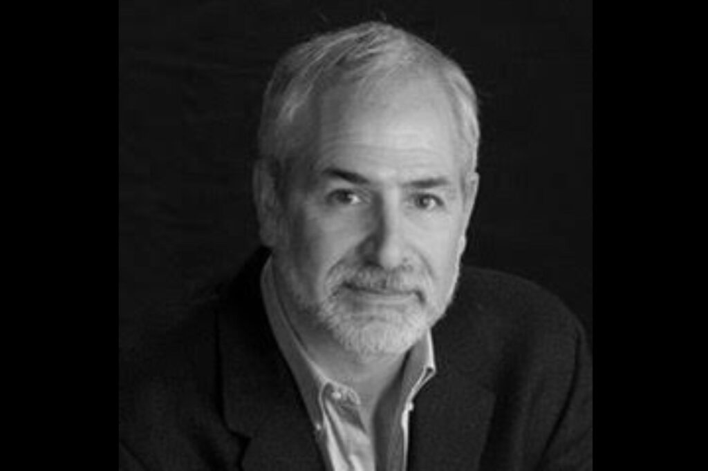 A grayscale photo of an older man with short hair and a beard, wearing a dark suit jacket over a light shirt. He gazes directly at the camera with a neutral expression, as if contemplating the vital work of a food bank, all against a plain dark background.