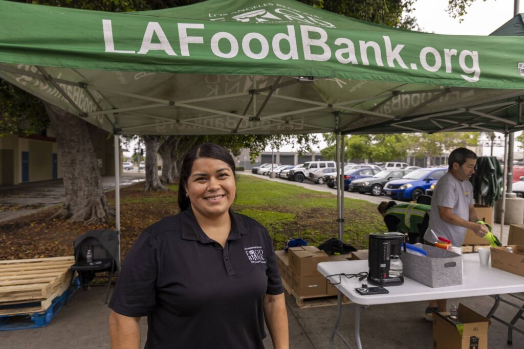 A woman in a black polo shirt smiles under a green LAFoodBank.org tent, while another person organizes food pantry supplies on a table outside, with parked cars visible in the background.