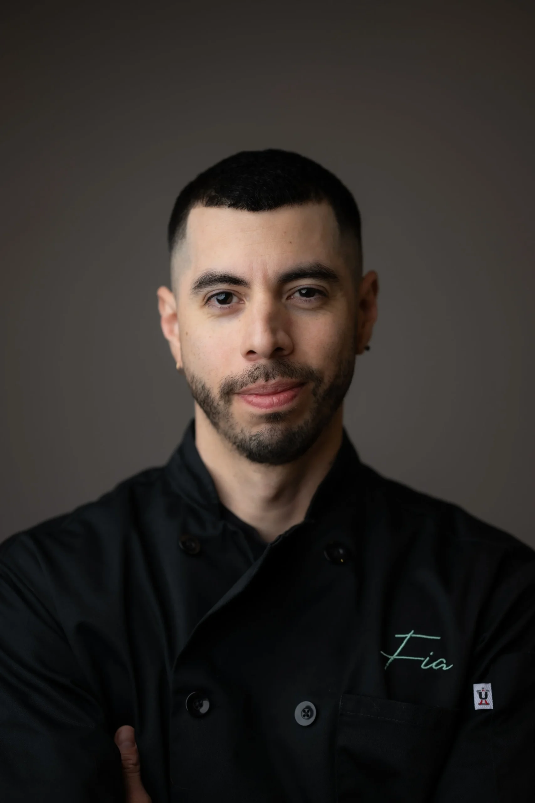 2025-04-17-FIA_portrait-017 A man with short dark hair and a trimmed beard wears a black chef's coat embroidered with the name "Fia," standing against a plain, dark background.