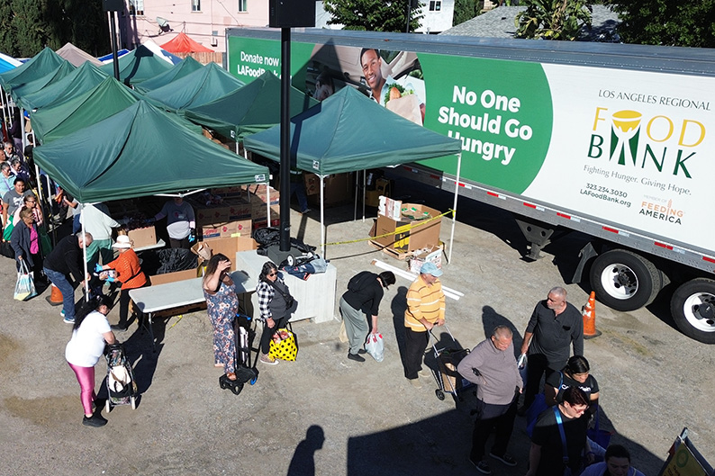 People wait in line at an outdoor WeFeedLA food distribution event next to a truck labeled 