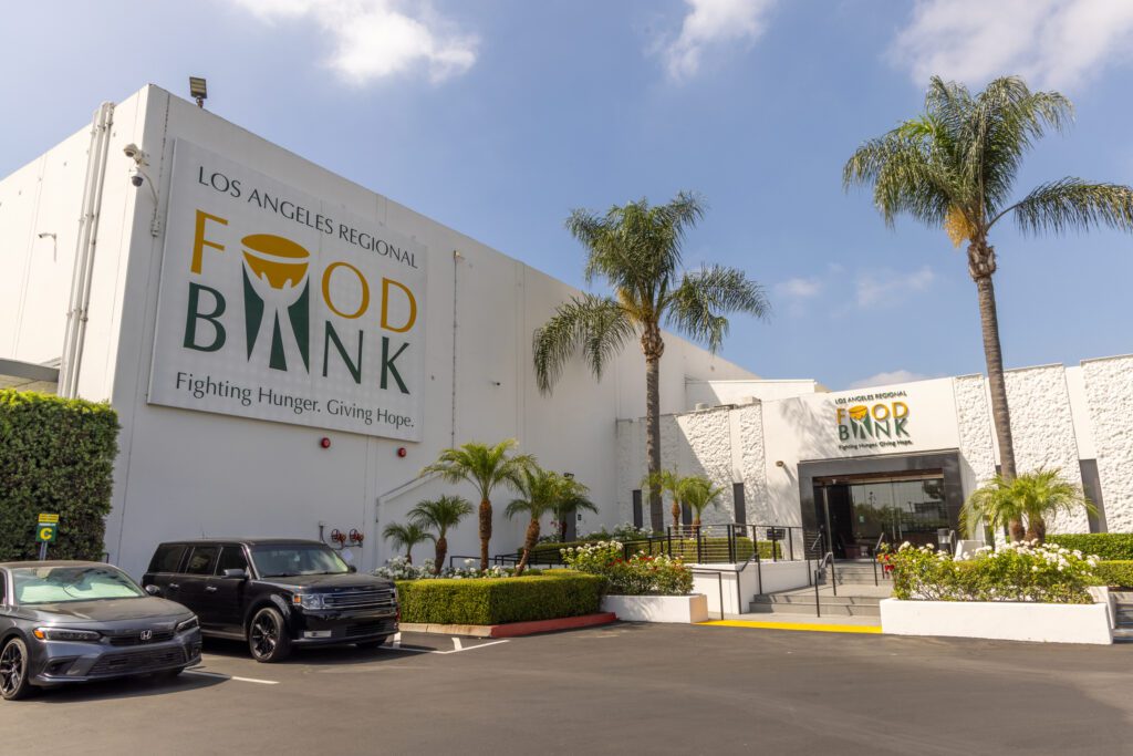 The exterior of the Los Angeles Regional Food Bank building, with large signage, palm trees, parked cars, and a clear blue sky above, invites visitors for a tour.