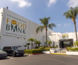 The exterior of the Los Angeles Regional Food Bank building, with large signage, palm trees, parked cars, and a clear blue sky above, invites visitors for a tour.
