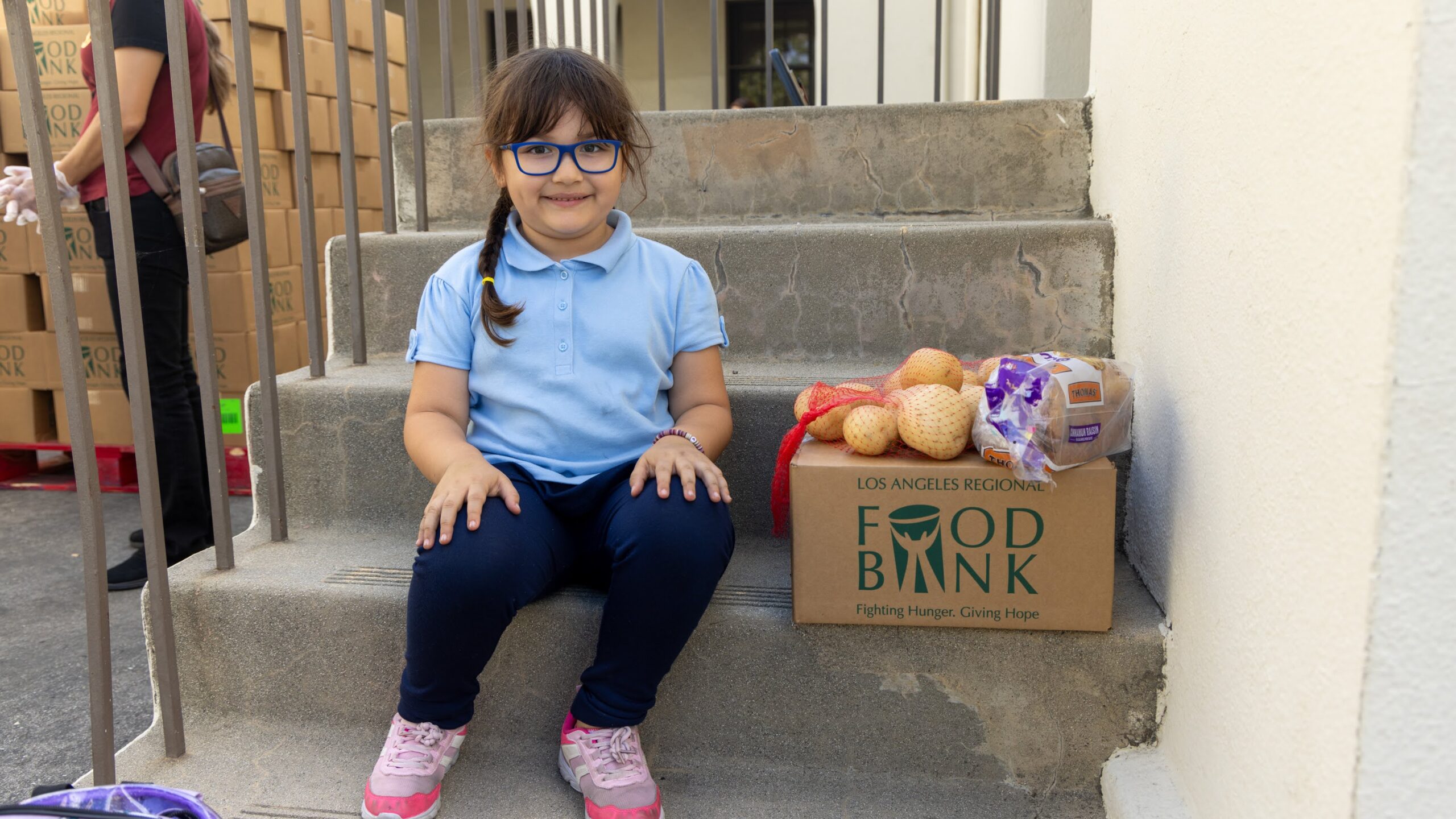 A young person sits on outdoor steps next to a cardboard box labeled "Los Angeles Regional Food Bank," containing groceries like onions and bread. She is smiling, wearing glasses, a light blue shirt, dark pants, and pink sneakers.