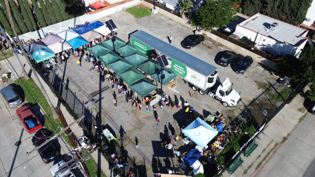 Aerial view of a Food Bank event with green and blue tents, a large white truck, and crowds gathered on a paved lot surrounded by fences and parked cars.