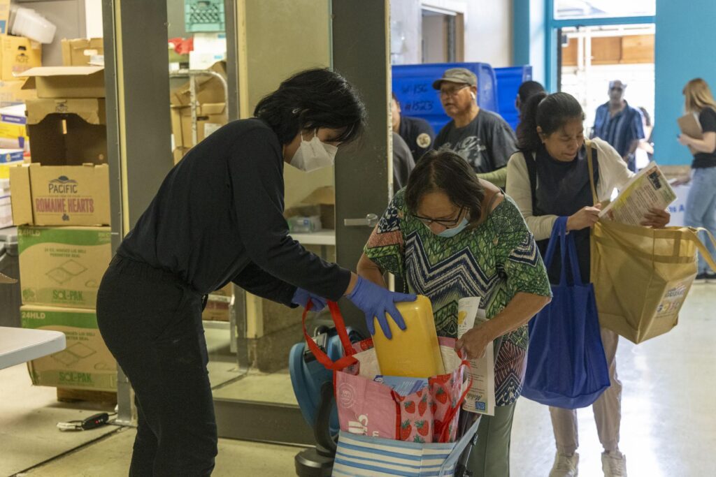 A volunteer wearing gloves and a mask helps a woman pack groceries and a food box into bags at a community food distribution event, while others wait in line holding bags.