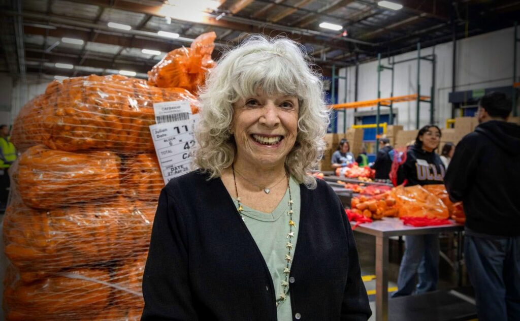 Roberta Stone smiles in the Los Angeles Regional Food Bank's warehouse, standing before large bags of oranges. In the background, people sort produce on tables.