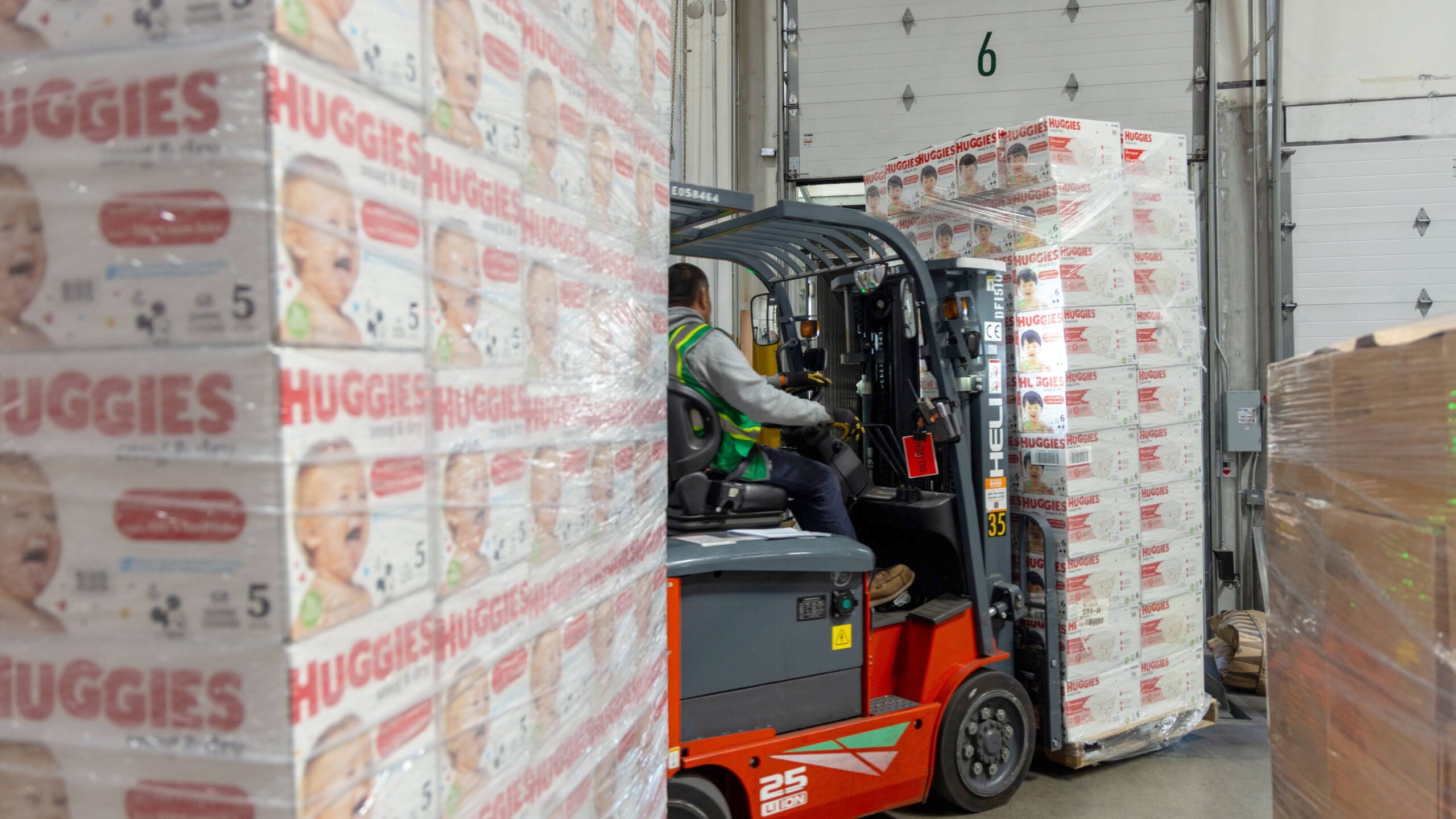 A worker operates a forklift to move pallets stacked with boxes of Huggies diapers, part of a diaper assistance program, inside a warehouse. The boxes feature images of smiling babies.