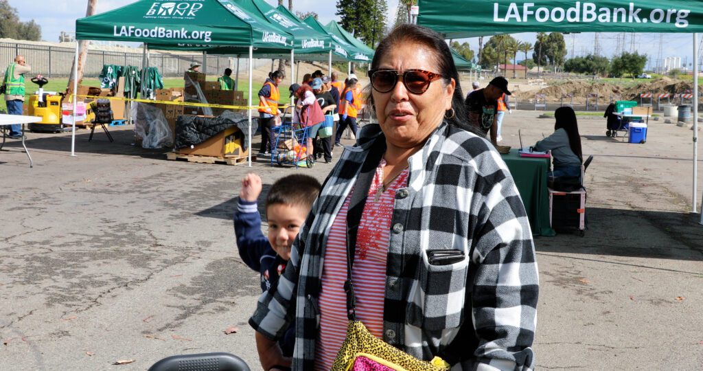 A woman in sunglasses and a plaid jacket stands outdoors with a child beside her at a mobile food pantry event, with LA Food Bank tents and volunteers visible in the background.