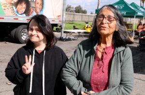 Two women stand outdoors by a tent and truck, possibly at a mobile food pantry. One woman with Down syndrome in a black hoodie shows a peace sign, while the older woman in glasses and a green jacket smiles beside her.
