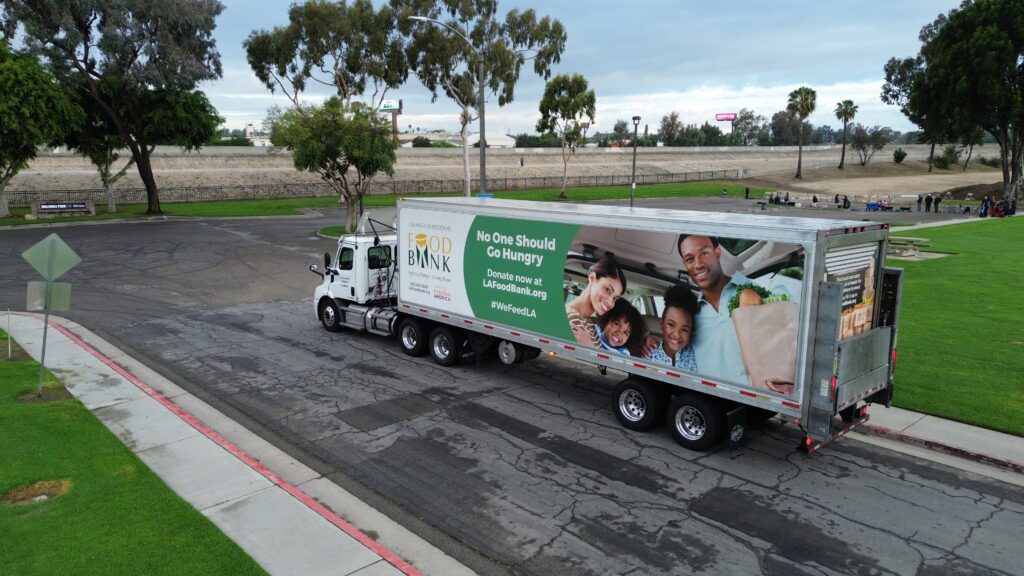 A large truck with a “Food Bank” sign and a photo of a smiling family serves as a mobile food pantry, carrying groceries. The truck is parked on a paved road in a park, surrounded by trees and grass.